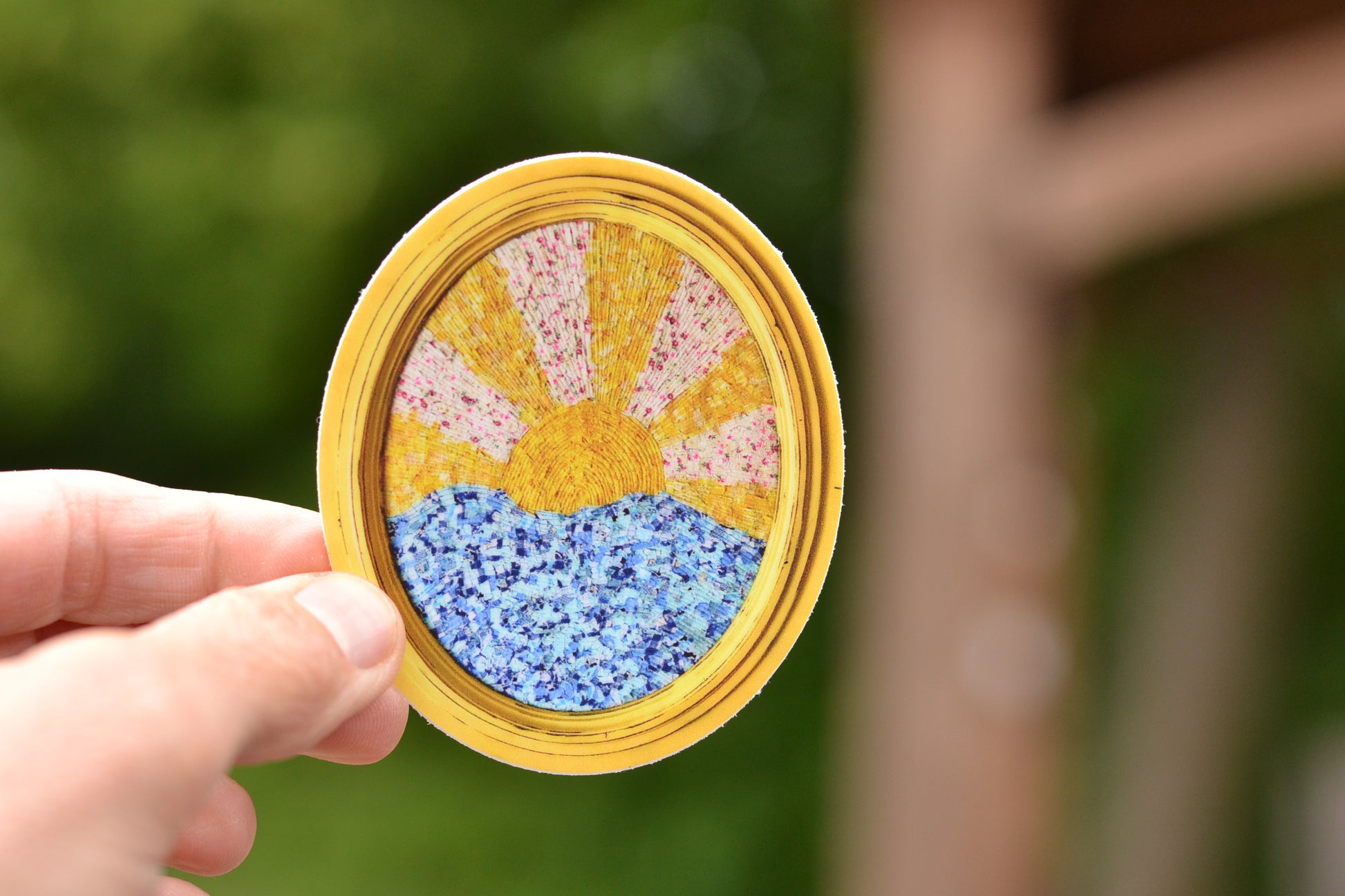 Hand holding a decorative jar with a sun design against a blurred green background